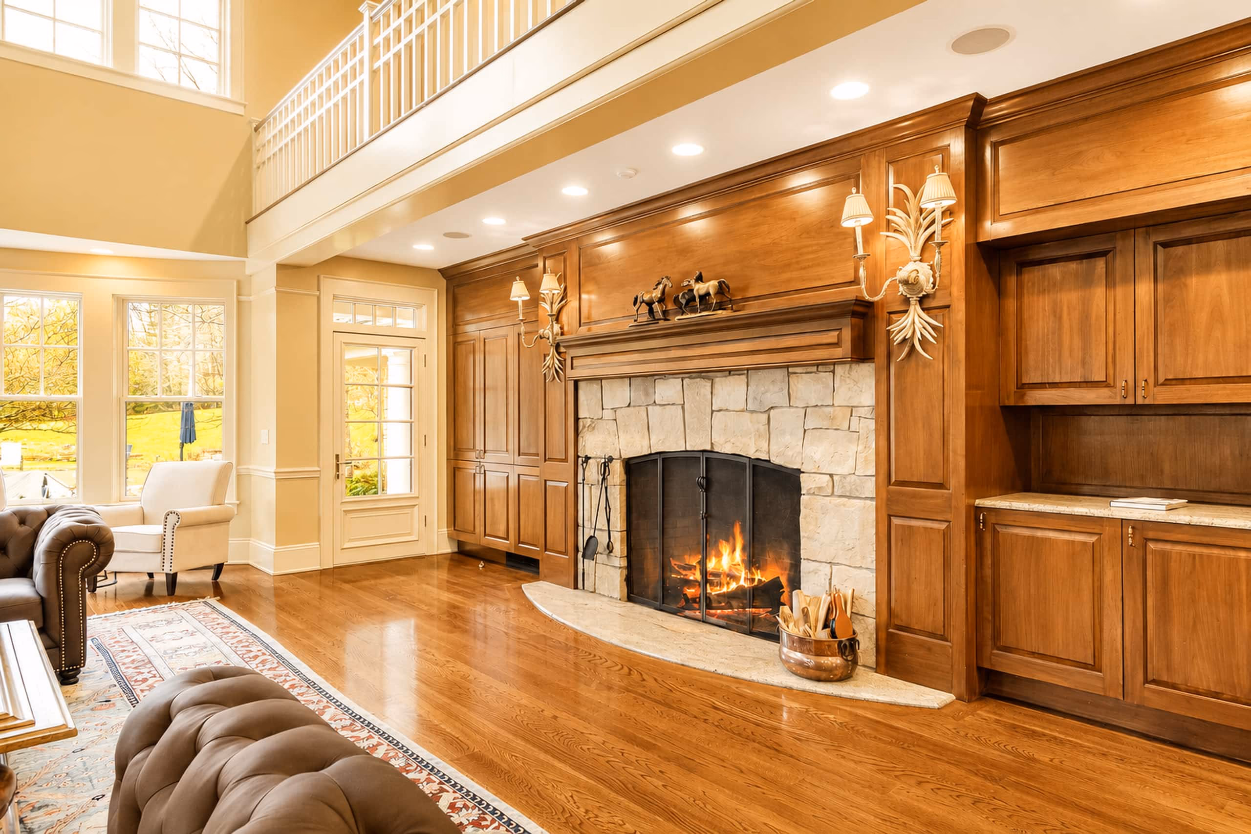 Interior view of the Great Room focusing on the massive stone fireplace with a fire crackling inside the hearth. Framed by custom dark wood paneling, columns, and built-in display shelves. The room has shiny hardwood floors. [Season: Indoor] [Lighting: Warm, cozy firelight and soft recessed spotlights] [Content: Stone fireplace, fireplace hearth, wood paneling, great room details]