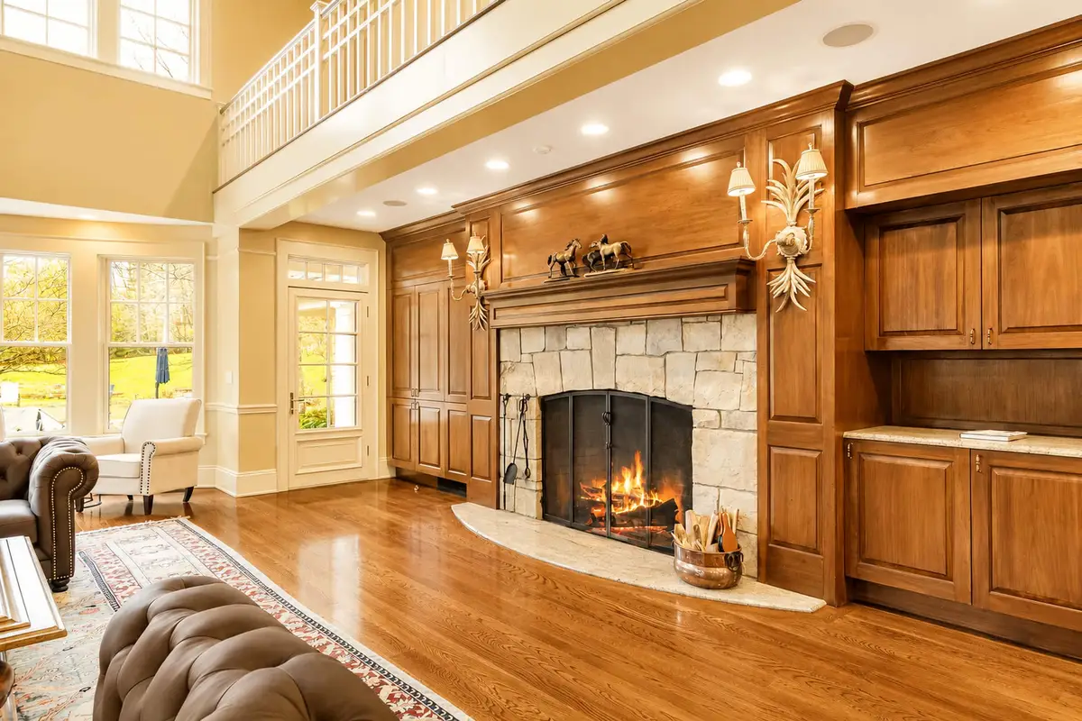 Interior view of the Great Room focusing on the massive stone fireplace with a fire crackling inside the hearth. Framed by custom dark wood paneling, columns, and built-in display shelves. The room has shiny hardwood floors. [Season: Indoor] [Lighting: Warm, cozy firelight and soft recessed spotlights] [Content: Stone fireplace, fireplace hearth, wood paneling, great room details]