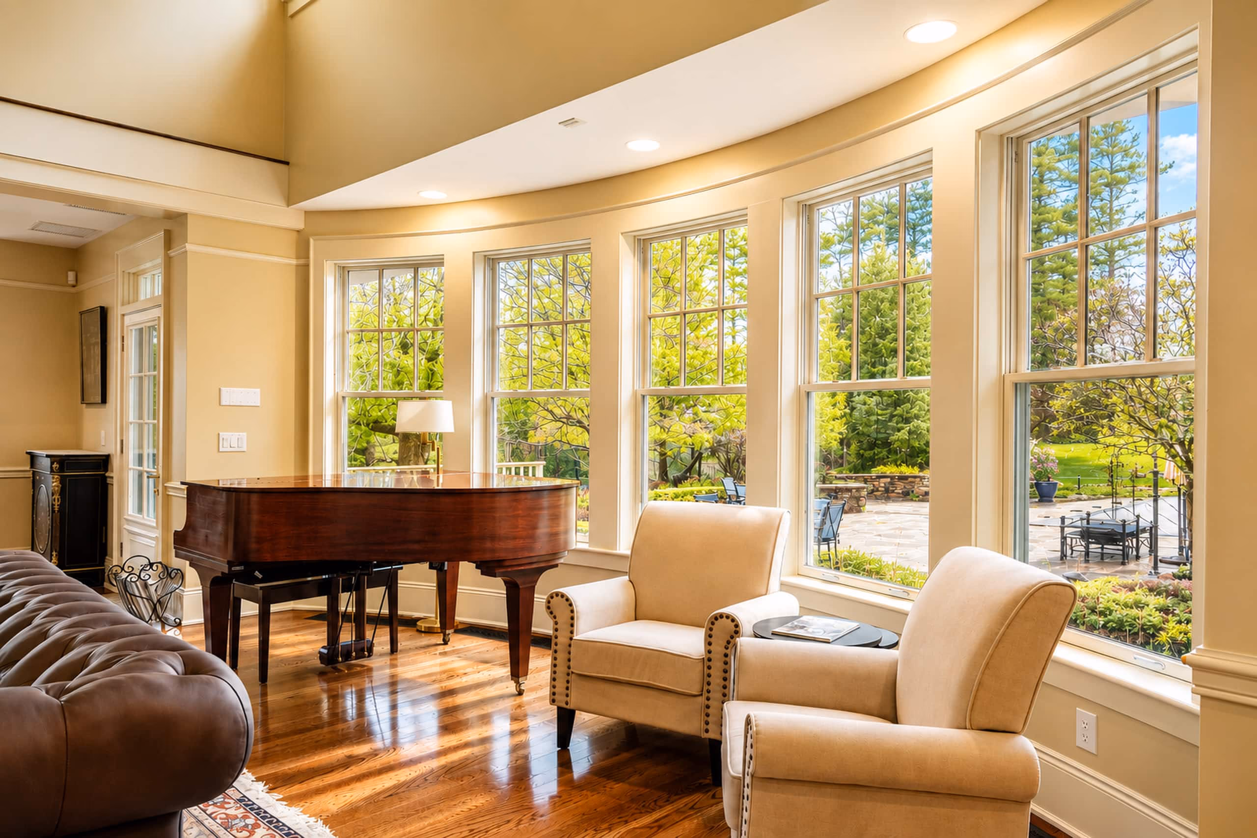 Close-up of the sunlit east-facing conversation seating area in the Great Room. Features two plush beige armchairs sitting next to a mahogany grand piano in front of a bank of large, tall double-hung windows looking out onto lush green lawns and trees. [Season: Summer] [Lighting: Bright, warm afternoon daylight] [Content: Great room seating, beige armchairs, grand piano, window view]