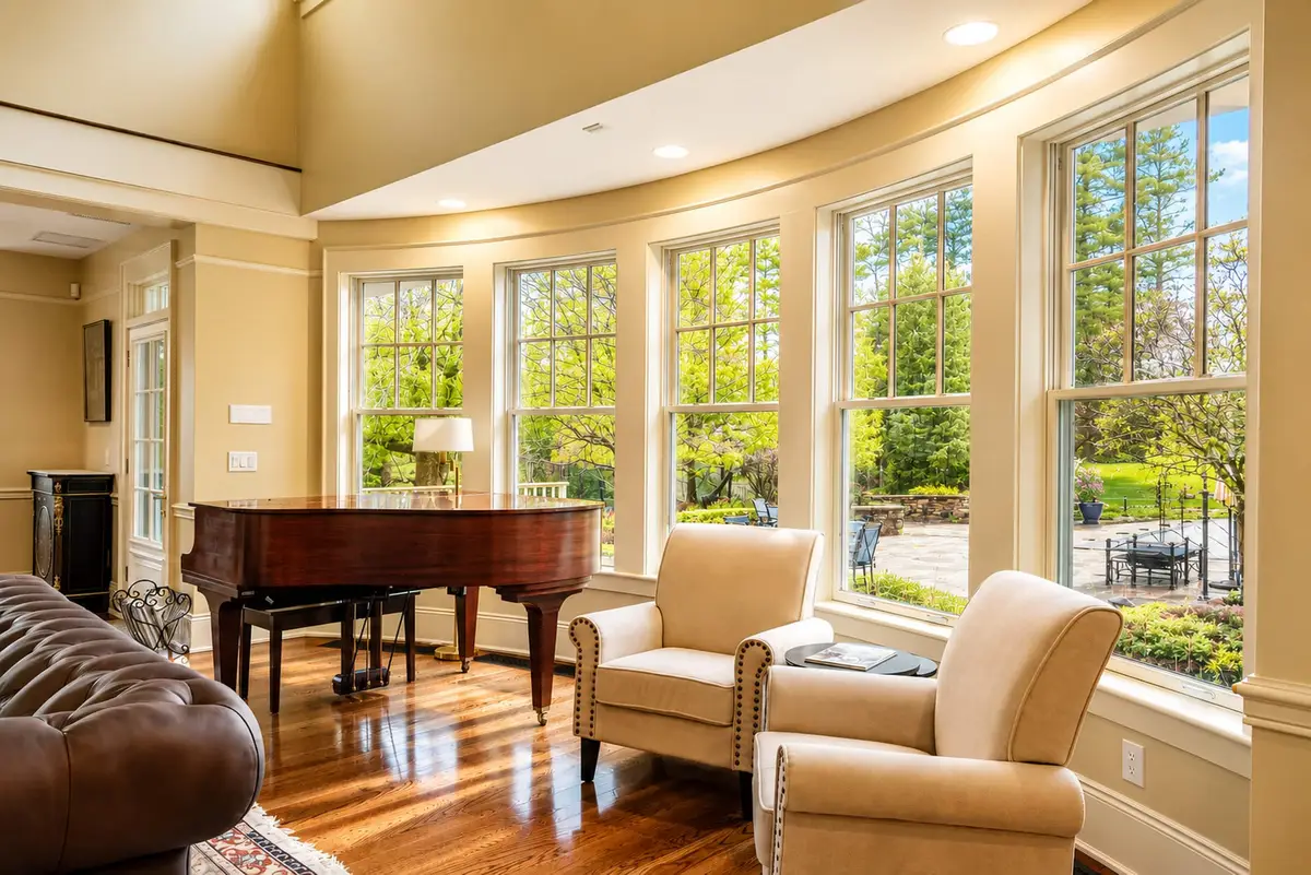 Close-up of the sunlit east-facing conversation seating area in the Great Room. Features two plush beige armchairs sitting next to a mahogany grand piano in front of a bank of large, tall double-hung windows looking out onto lush green lawns and trees. [Season: Summer] [Lighting: Bright, warm afternoon daylight] [Content: Great room seating, beige armchairs, grand piano, window view]