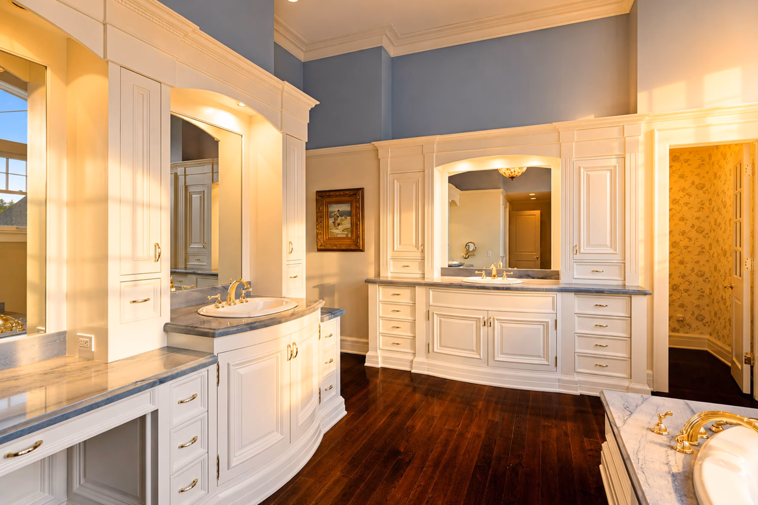 Master bathroom vanity showing custom white cabinets, a dark wood floor, a double marble vanity countertop with undermount sinks, gold faucets, and large mirrored alcoves under soft vanity lights. [Season: Indoor] [Lighting: Warm, clean vanity lighting] [Content: Double vanity, white cabinets, marble counter, bathroom mirrors]