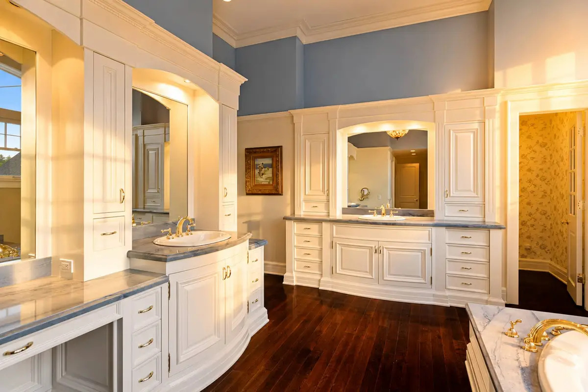 Master bathroom vanity showing custom white cabinets, a dark wood floor, a double marble vanity countertop with undermount sinks, gold faucets, and large mirrored alcoves under soft vanity lights. [Season: Indoor] [Lighting: Warm, clean vanity lighting] [Content: Double vanity, white cabinets, marble counter, bathroom mirrors]