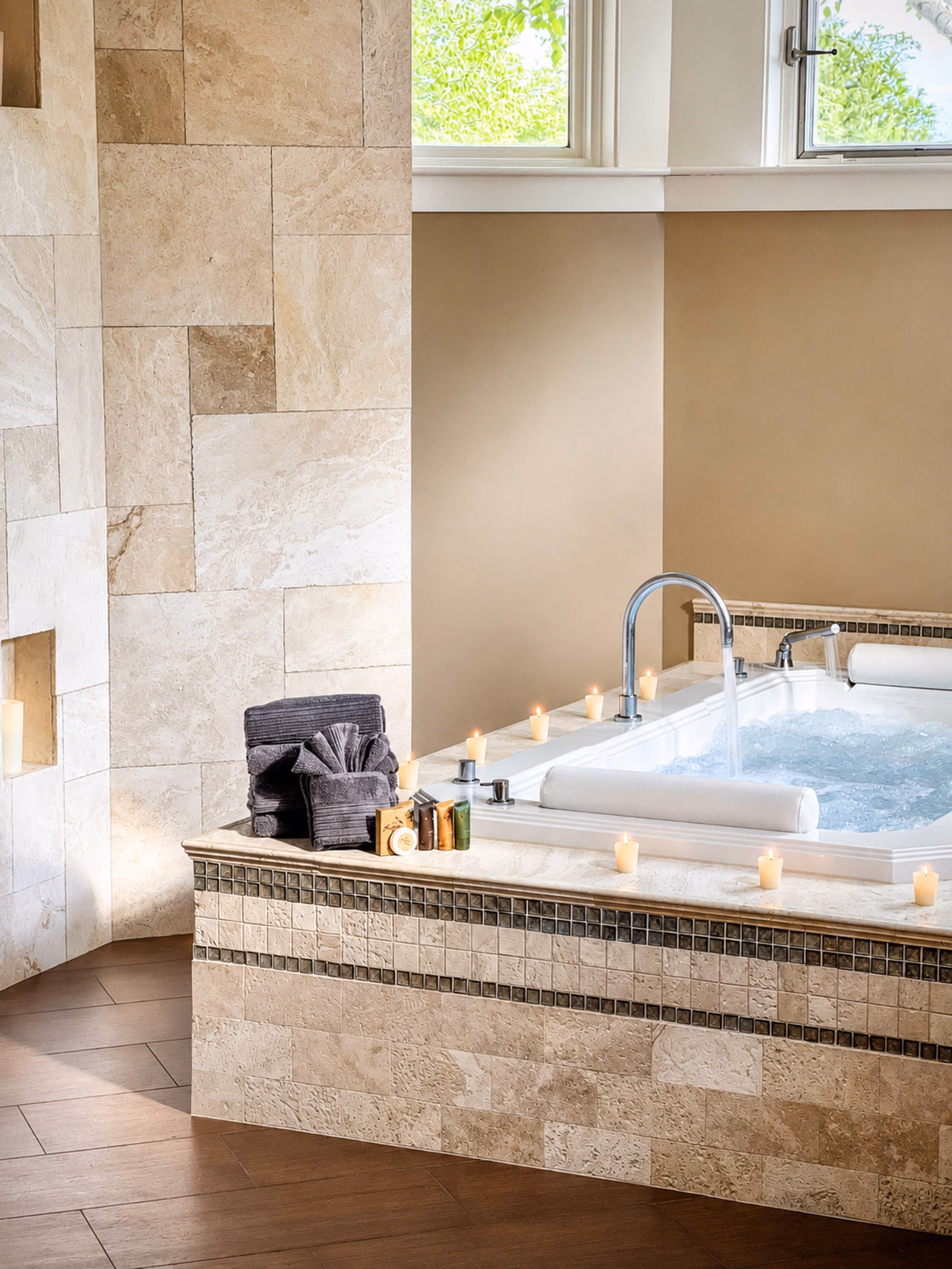 Indoor spa area showing a large white soaking tub set in a light marble tile surround with candles lit along the deck. A grey rolled towel sits on the edge. [Season: Indoor] [Lighting: Soft, warm, relaxing candle-lit and ambient light] [Content: Spa soaking tub, marble tiling, lit candles, relaxation area]