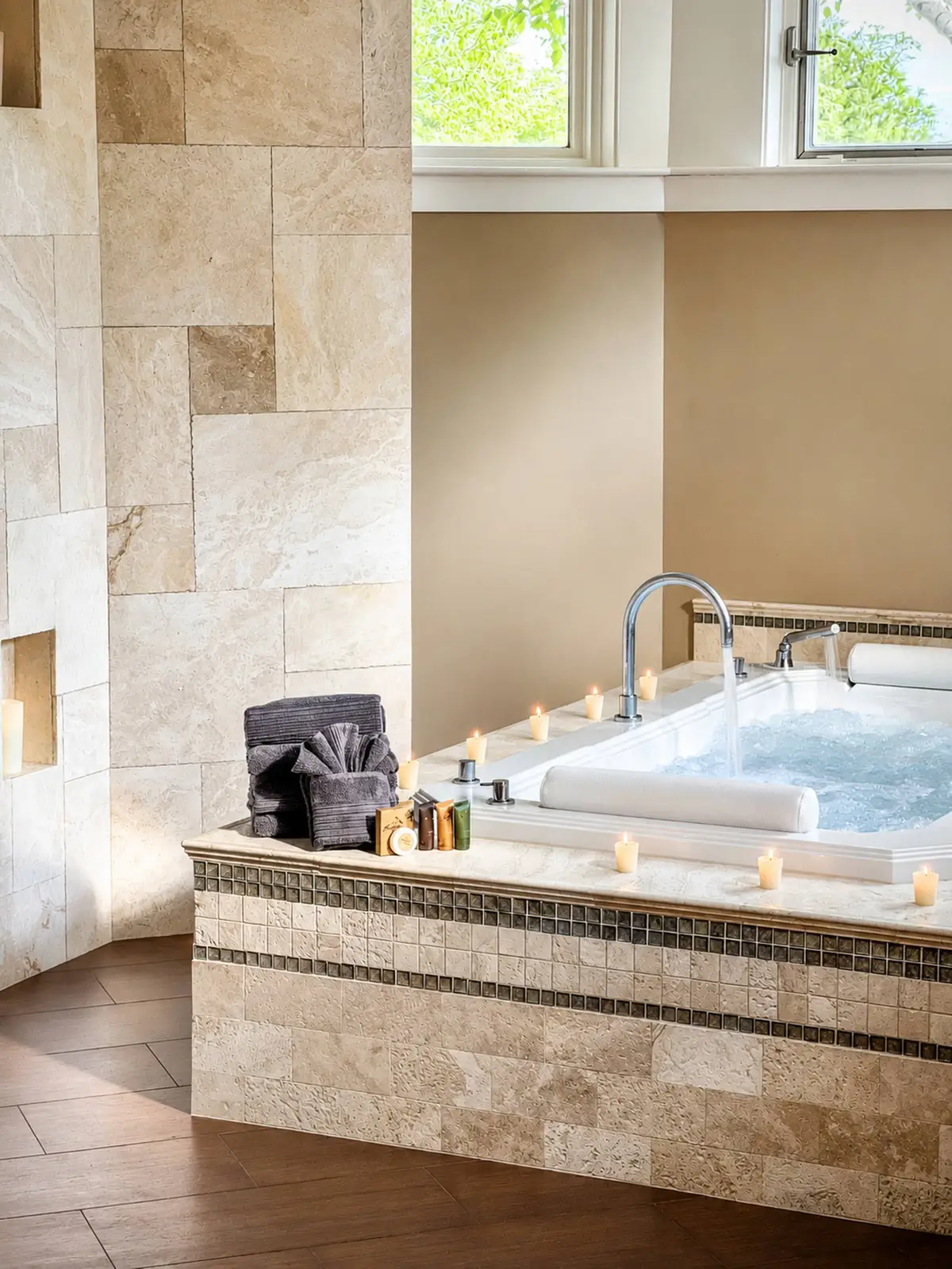 Indoor spa area showing a large white soaking tub set in a light marble tile surround with candles lit along the deck. A grey rolled towel sits on the edge. [Season: Indoor] [Lighting: Soft, warm, relaxing candle-lit and ambient light] [Content: Spa soaking tub, marble tiling, lit candles, relaxation area]