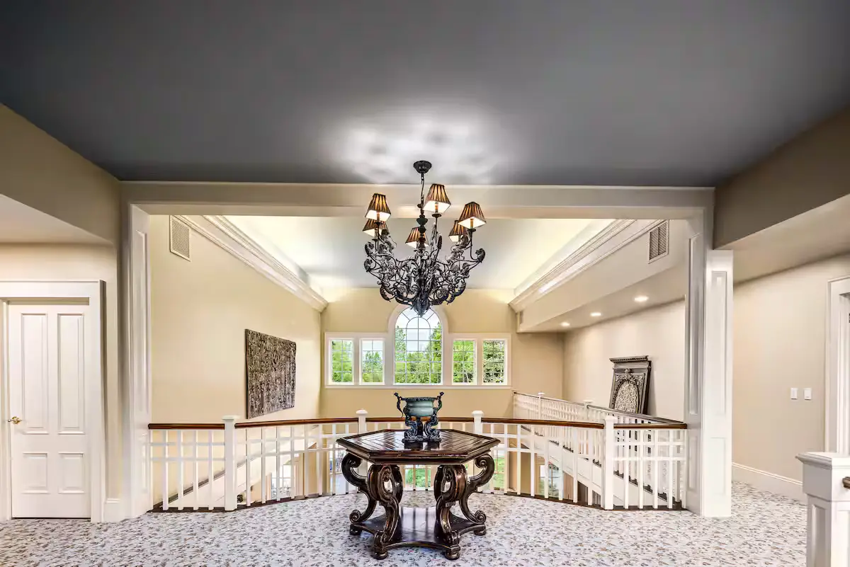 Wide-angle interior view of the upper-level loft landing. Features an ornate dark wrought-iron chandelier with small lampshades hanging from a dark blue-grey ceiling, casting decorative shadows. Below it sits a dark wood octagonal table with scrolled legs on a floral-patterned carpet. White balustrades overlook the lower level, and windows look out on green summer trees. [Season: Summer] [Lighting: Combined warm chandelier and natural daylight] [Content: Loft landing, hallway, chandelier, antique table, stairs overview]