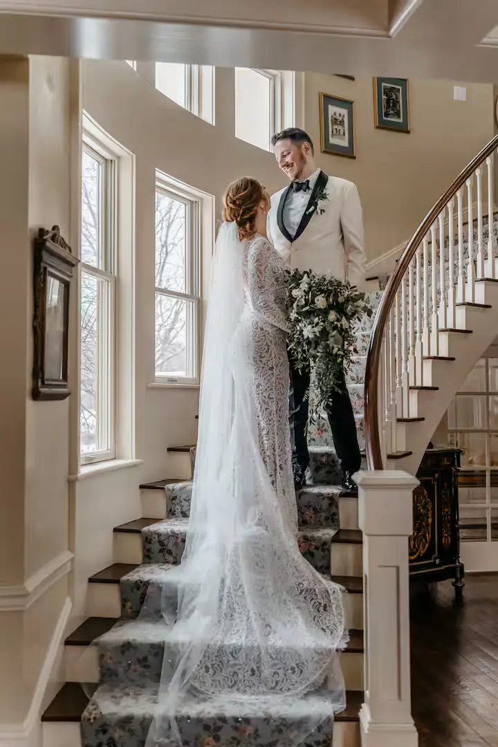 Indoor portrait-style shot of a bride in a white lace wedding gown holding a floral bouquet, standing on a staircase next to a groom in a dark tuxedo. A large window and white wooden railings are visible in the background, creating a romantic and elegant setting. [Season: Summer/Spring] [Lighting: Soft, diffused natural daylight] [Content: Wedding couple, staircase, formal attire, indoor portrait]