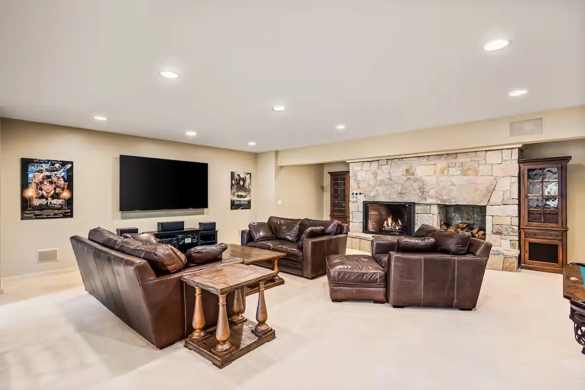 Wide view of a spacious downstairs family and game room. Features a conversation area with two large brown leather sofas and a matching armchair on a light carpet around wood and stone tables. In the background is a flat-screen TV on the wall, a fireplace with a lit fire, and warm wood-paneled walls. [Season: Autumn/Winter] [Lighting: Soft, warm recessed ceiling lights and fire glow] [Content: Family room, leather furniture, fireplace, media room interior]