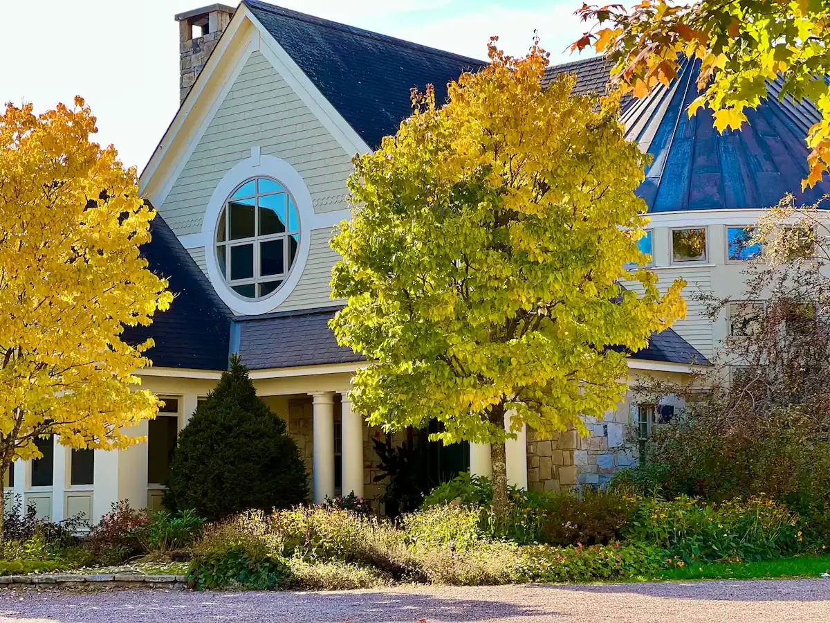 Exterior daytime view of the estate's grand front entrance. Features stone columns, robust timber framing, a round architectural window, and a unique circular copper-domed wing. Bright autumn foliage with yellow-orange leaves frames the house under a clear, bright blue sky with sharp daylight casting distinct shadows. [Season: Autumn] [Lighting: Bright, direct daylight] [Content: Front elevation, circular driveway, landscaping, stone masonry, architectural exterior]