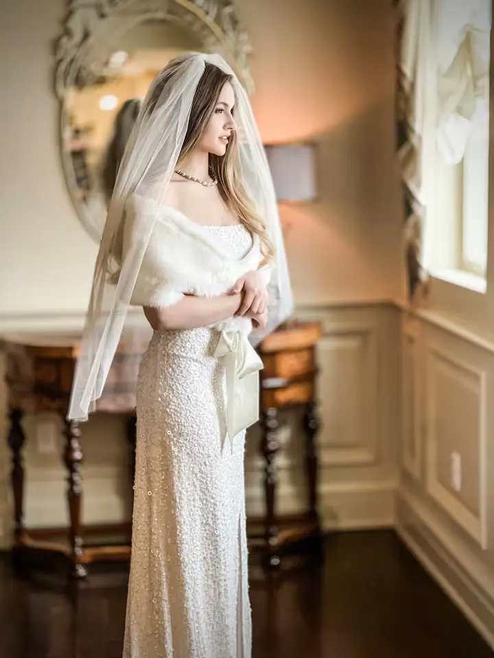 Indoor shot of a bride in a white wedding dress and a white fur shoulder wrap, standing next to a wooden console table. The background features light beige walls with elegant trim. [Season: Indoor] [Lighting: Soft, warm indoor lighting] [Content: Bride portrait, formal dress, hallway interior]