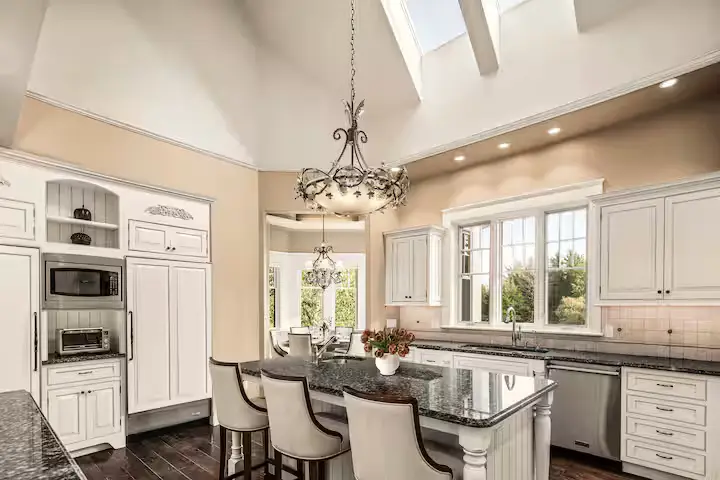 Wide-angle view of the gourmet main kitchen showcasing high vaulted ceilings, light beige walls, a circular iron chandelier, custom white cabinets, dark granite countertops, and a central island with high-backed bar chairs. Outside the window behind the sink, green trees are visible. [Season: Summer/Spring] [Lighting: Bright, natural daylight] [Content: Gourmet kitchen, central island, cabinetry, high ceilings, kitchen interior]