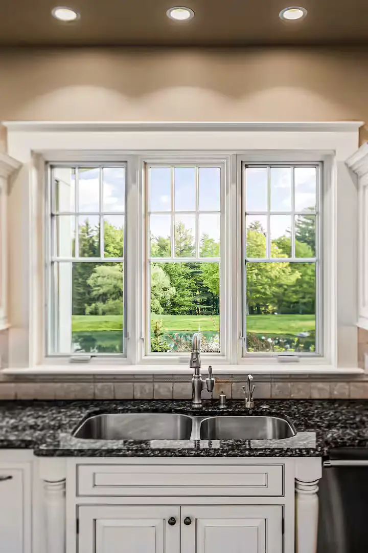 Close-up view of a kitchen double-basin undermount sink with a gooseneck faucet, set in dark granite countertops directly under a large three-pane window that looks out at a lush green backyard lawn and a calm pond. [Season: Summer/Spring] [Lighting: Bright daylight pouring in] [Content: Kitchen sink, granite counter, window view, backyard pond]