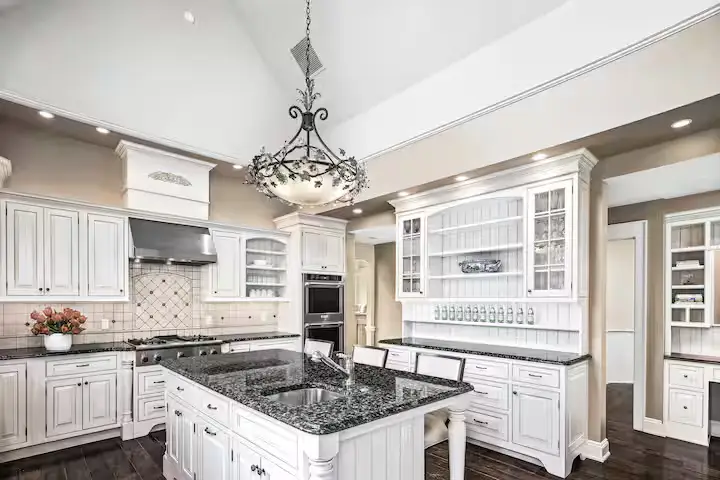 Wide-angle perspective of the gourmet main kitchen. Shows custom white-painted cabinets, a large central island with a dark granite countertop and breakfast bar seating, double built-in ovens, a massive range hood, and a crystal chandelier hanging from the high tray ceiling. [Season: Summer/Spring] [Lighting: Bright, polished daytime light] [Content: Gourmet kitchen, white cabinets, island seating, granite countertops, kitchen interior]