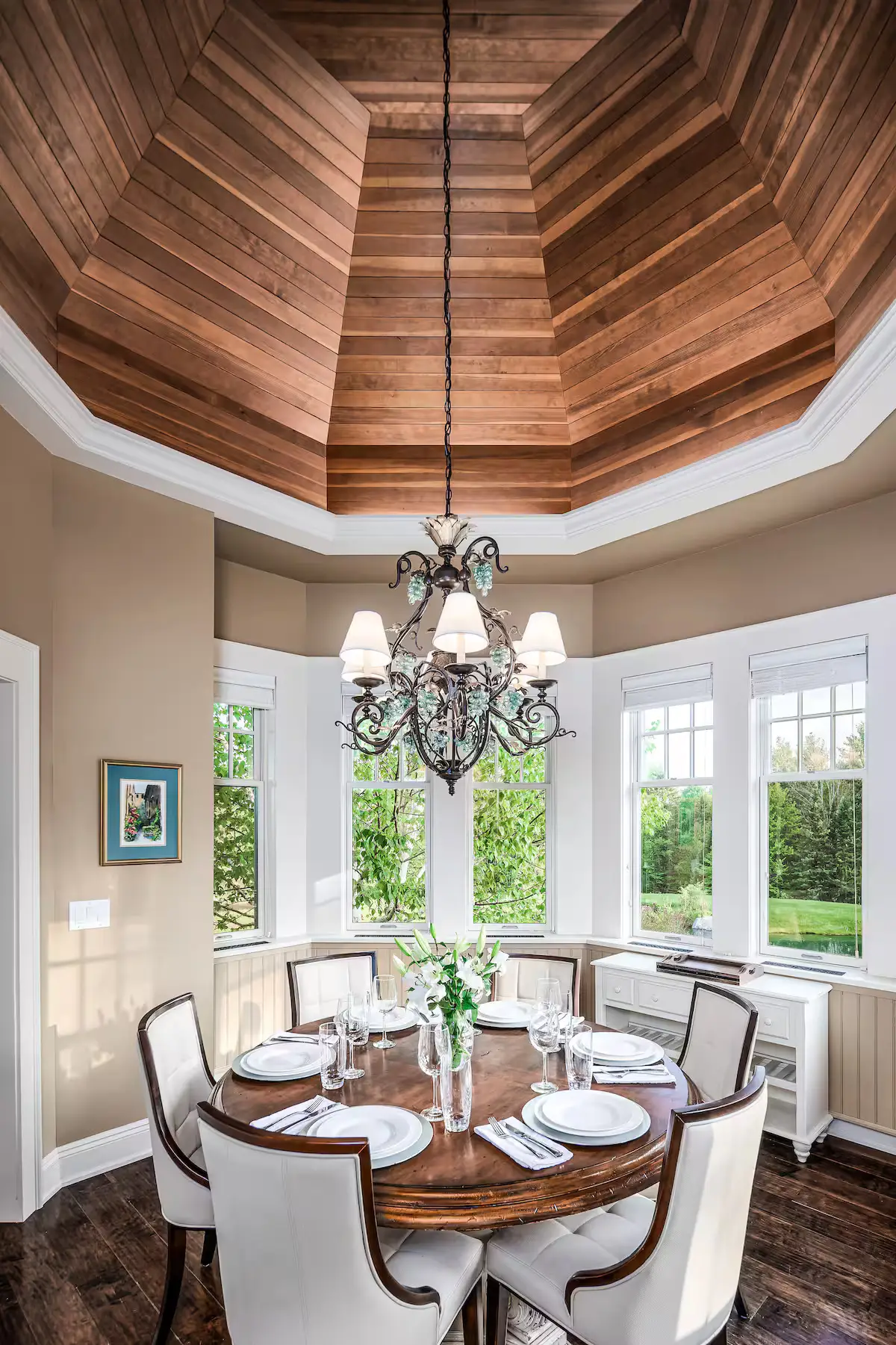 Casual dining or sunroom nook set inside a multi-window bay alcove under a high octagonal wood-paneled ceiling with a detailed chandelier. Looking out to vibrant green summer foliage. [Season: Summer] [Lighting: Soft, diffused daylight] [Content: Breakfast nook, bay windows, wood ceiling, garden view]
