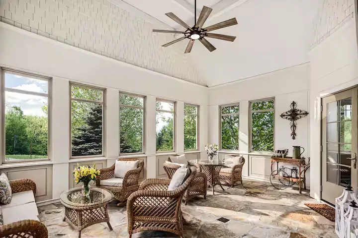 Spacious sunroom featuring custom wicker furniture (a sofa, armchairs, and a round coffee table), stone-tiled floor, a ceiling fan, and a line of tall windows looking out at green lawns and trees. [Season: Summer] [Lighting: Bright, direct daylight] [Content: Sunroom, wicker furniture, stone tile floor, garden view exterior]