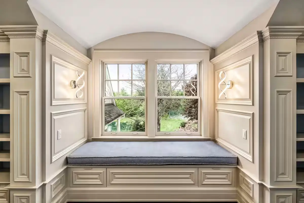 A sunlit window seat alcove in a guest bedroom. Features a blue cushion on the built-in bench, white trim paneling, and two wall sconces on either side. The window looks out onto green summer trees. [Season: Summer] [Lighting: Bright, soft daylight] [Content: Window seat, bedroom alcove, wall sconces, reading nook]