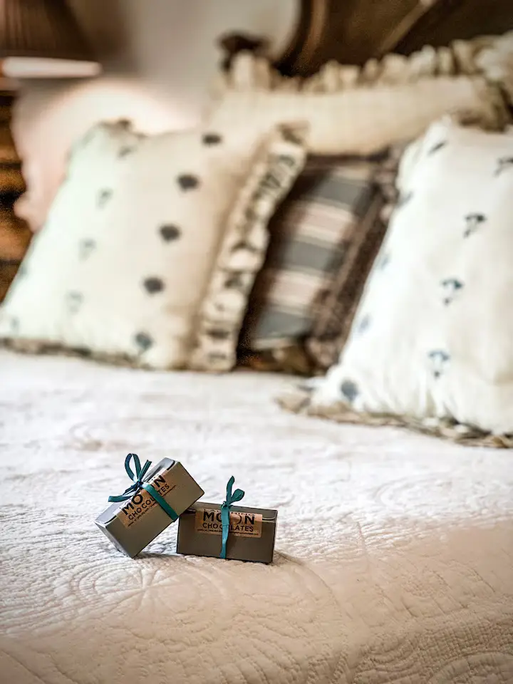 Close-up detail of a guest bed showing premium light-patterned pillows and a small box of 'MOUNT MANSFIELD CHOCOLATES' wrapped in a bow sitting on the duvet. [Season: Winter/Autumn] [Lighting: Warm, soft-focus macro lighting] [Content: Bed detail, pillows, welcome gift chocolates]