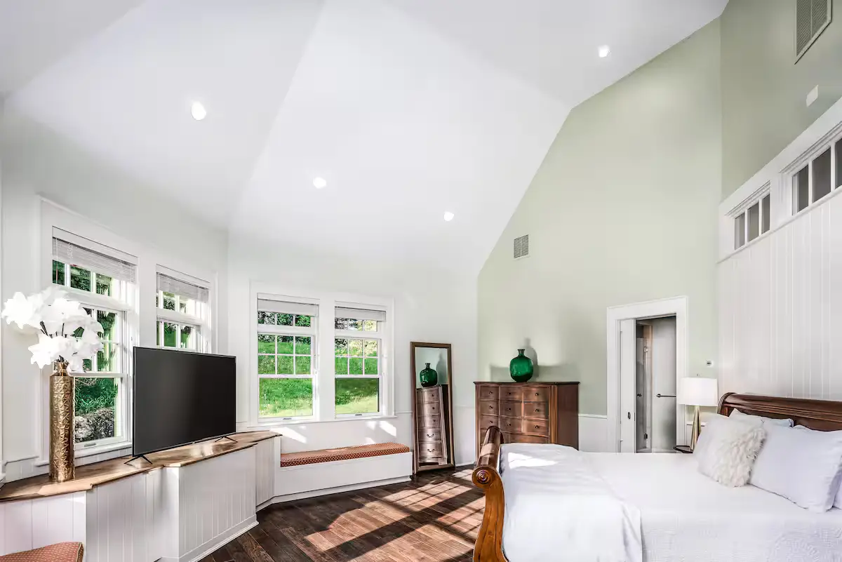 Spacious guest bedroom with a vaulted ceiling, light green walls, a king-sized bed with wooden posts, a white TV stand console, and bright windows looking out onto a green summer landscape. [Season: Summer] [Lighting: Bright daylight with distinct sun beams casting across the floor] [Content: Guest bedroom, king bed, vaulted ceiling, sunbeams]