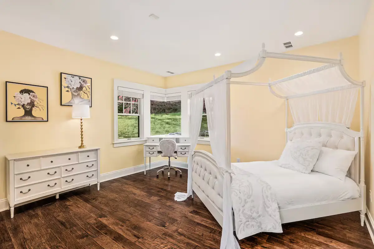 Whimsical bedroom featuring a white metal canopy four-poster queen bed, yellow walls, dark hardwood floors, a white dresser, and a window with green summer lawn views. [Season: Summer] [Lighting: Soft daylight and warm lamp light] [Content: Canopy bed, yellow bedroom, wood floors, garden view]