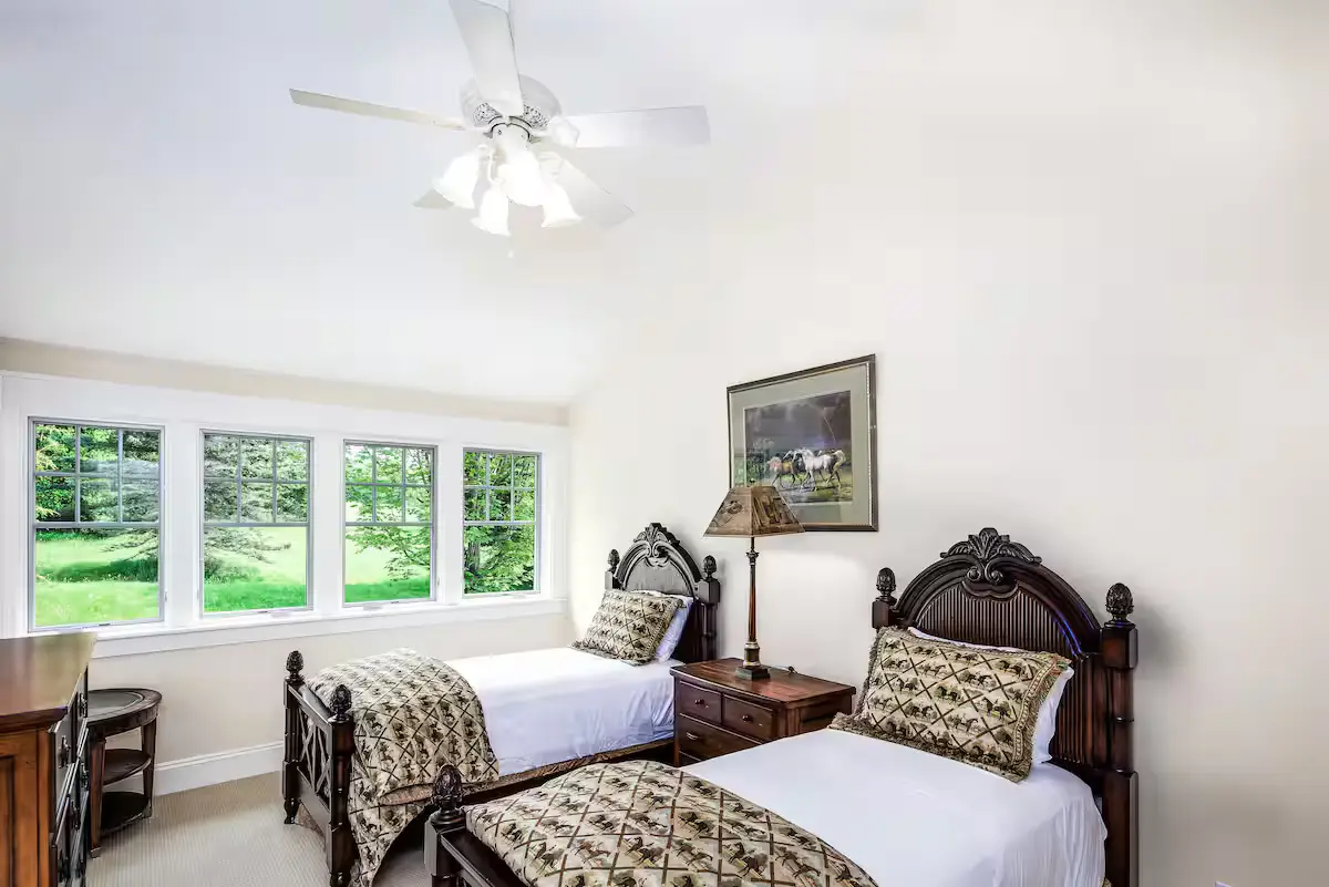 Charming guest bedroom with two twin beds featuring dark wood headboards and patterned gold/brown quilts. A ceiling fan, white walls, and a large three-pane window look out onto green grass. [Season: Summer] [Lighting: Bright, airy daylight] [Content: Twin bedroom, two beds, ceiling fan, garden view]