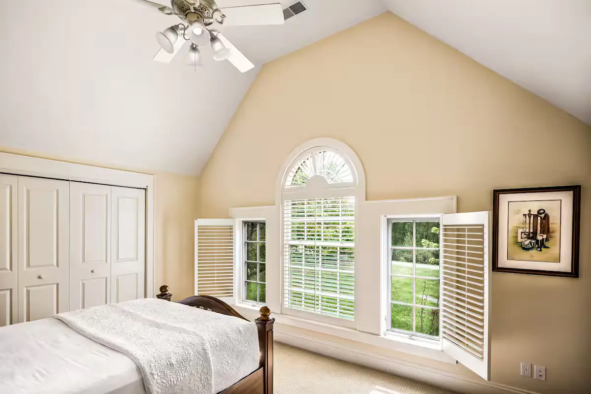 Guest bedroom under a vaulted ceiling with warm yellow-cream walls, featuring a bed with white linens, white bi-fold closet doors, a large central arched window flanked by two rectangular windows with open white shutters, showing green forest trees. [Season: Summer] [Lighting: Bright, diffused daytime light] [Content: Vaulted bedroom, white shutters, arched window, forest view]