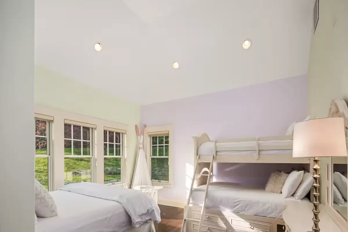 Children's or guest bedroom with a white bunk bed (twin over twin) and an additional twin bed, light purple-blue walls, and three windows overlooking a green field. [Season: Summer] [Lighting: Soft, pleasant daylight] [Content: Bunk beds, triple bedroom, kids room, field view]