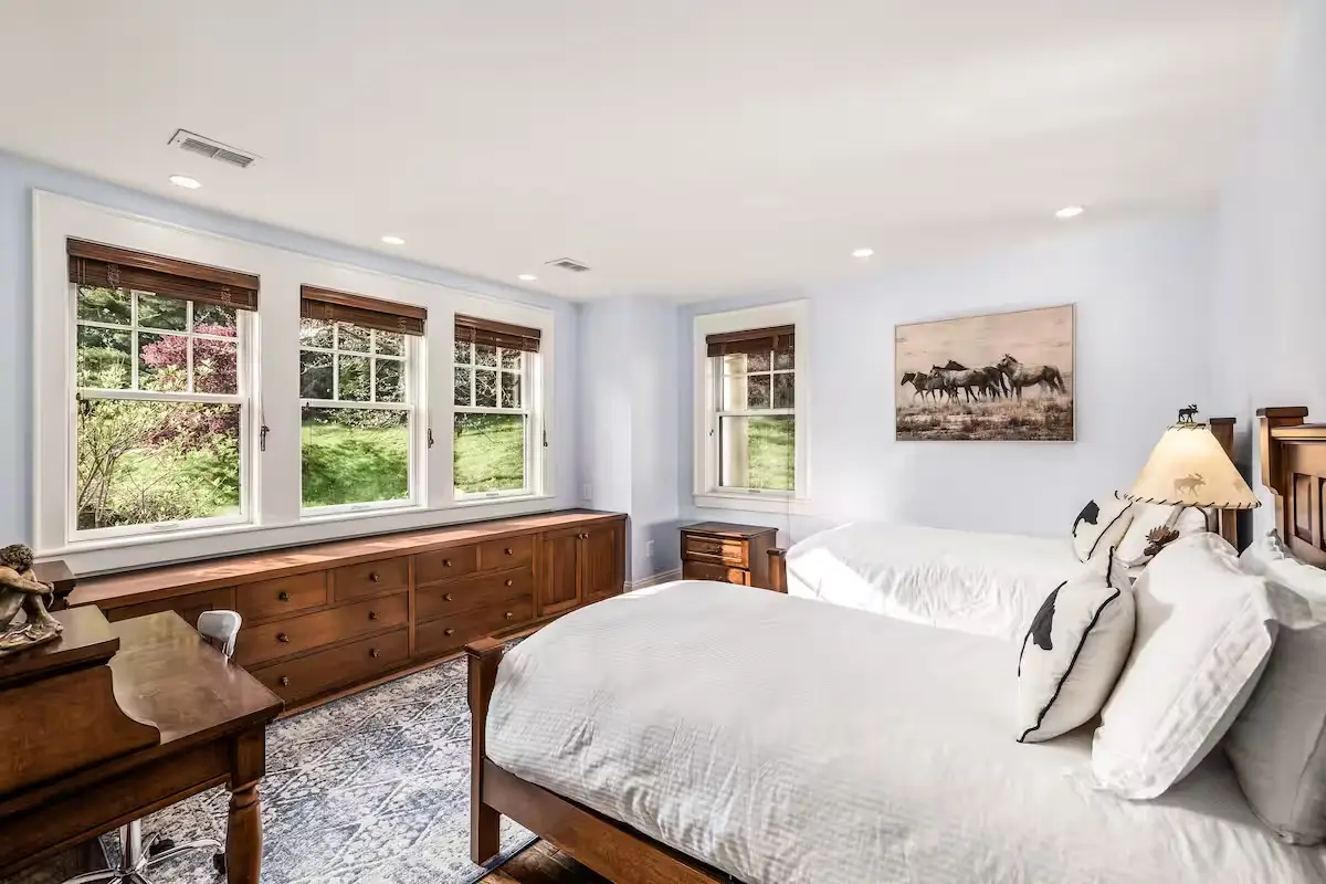 Guest bedroom with two queen beds dressed in white linens and decorative patterned throw pillows. Features a long dark wood storage console under three windows looking out at green summer lawns. An ornate rug is on the hardwood floor. [Season: Summer] [Lighting: Soft daylight and warm bedside lamp light] [Content: Double queen bedroom, wood console, lawn view]