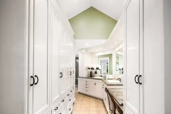 Pristine guest bathroom with a long white double-sink vanity, white custom cabinets on both sides, a marble countertop, large mirrors, a vaulted ceiling, and pale green walls. [Season: Indoor] [Lighting: Bright, clean vanity lighting] [Content: Guest bathroom, double vanity, white cabinets, vaulted ceiling]