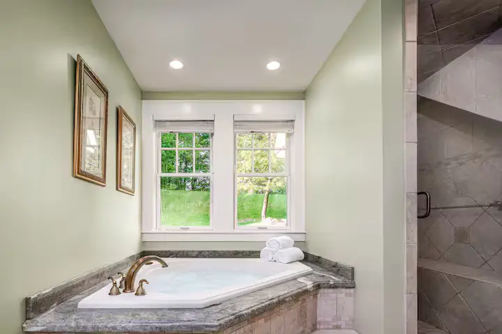Full bathroom ensuite featuring a white soaking tub set in a grey marble deck under two double-hung windows with white frames looking out onto green summer trees. The walls are painted a soft green, and two framed prints hang on the wall. [Season: Summer] [Lighting: Soft, bright daylight] [Content: Soaking tub, green bathroom, double windows, garden view]