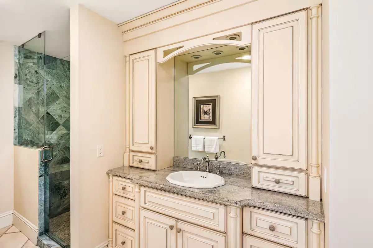 Full guest bathroom ensuite with a white double vanity, granite countertop, an undermount sink, classic white cabinets and columns framing a mirrored cabinet, and a walk-in shower on the left with green/grey marble tiling. [Season: Indoor] [Lighting: Warm, clean vanity light] [Content: Guest bathroom, white vanity, granite counter, marble shower]