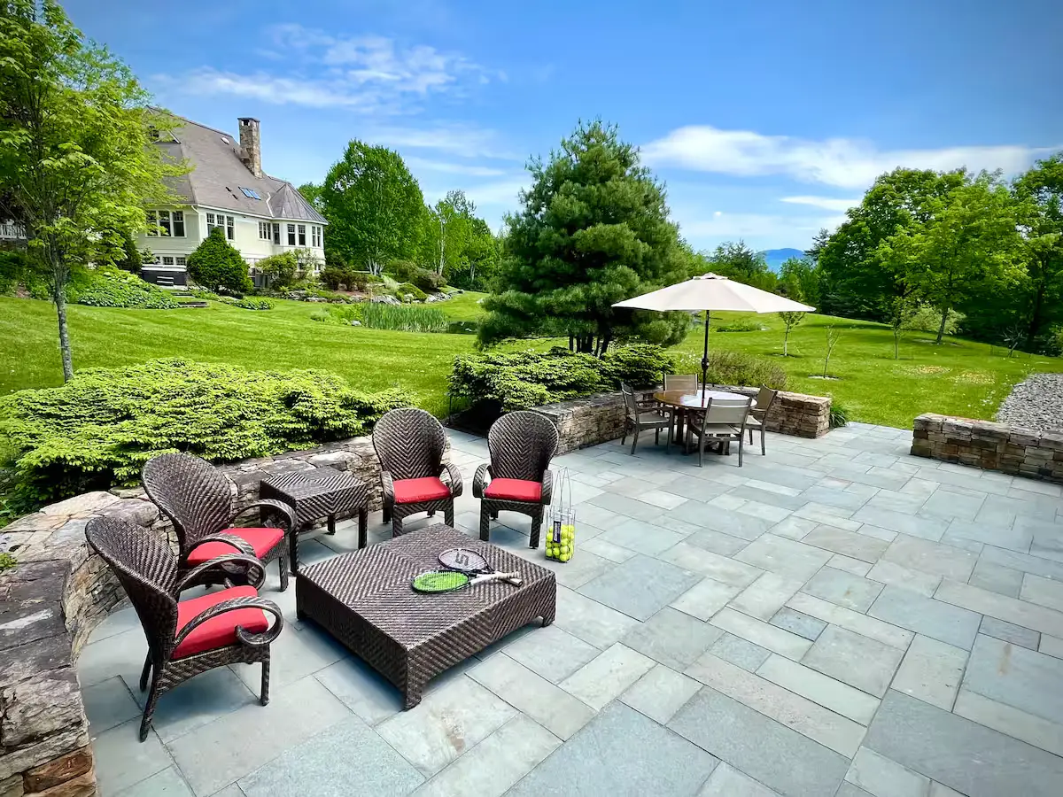 A luxurious stone-paved pool patio lounge set with premium dark wicker sofas and chairs featuring deep red cushions around a coffee table. Next to it is a dining table under a white umbrella, surrounded by manicured green lawns and trees. [Season: Summer] [Lighting: Soft afternoon daylight] [Content: Patio seating, pool deck, wicker sofas, umbrella table, backyard exterior]