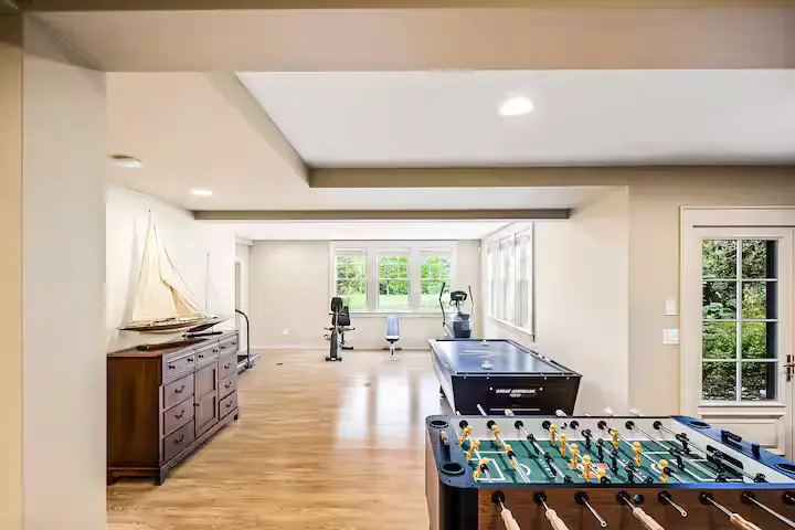 A spacious recreation and fitness room. Features a wooden foosball table in the foreground, polished hardwood floors, a wooden dresser with a decorative model sailboat on top, and fitness equipment (treadmills/elliptical) visible in the background near a window. [Season: Indoor] [Lighting: Bright, recessed ceiling lights] [Content: Recreation room, foosball table, fitness gear, wood floors]