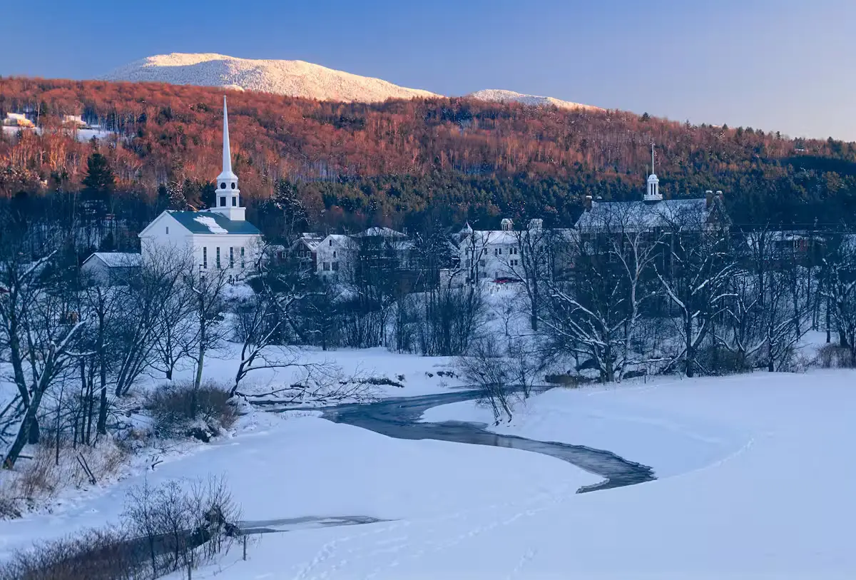 Panoramic winter landscape showing a winding, frozen river or stream in a snow-covered valley. A classic white church steeple and village buildings are nestled in the snow-covered forest. In the background are majestic mountains dusted in snow. [Season: Winter] [Lighting: Cool winter daylight] [Content: Winter village, snowy river, church steeple, mountain range]