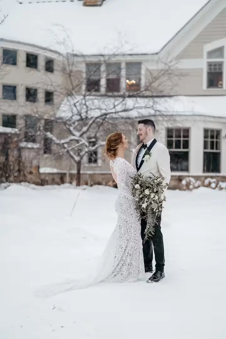 A bride and groom embracing and looking at each other in a snowy outdoor courtyard, with the estate house's stone facade visible in the background. The ground is blanketed in thick snow. [Season: Winter] [Lighting: Soft, diffused winter daylight] [Content: Wedding couple, snow courtyard, winter wedding, outdoor portrait]