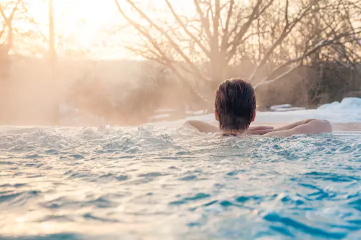 Close-up detail from behind a person soaking in an outdoor hot tub/spa, looking out over the water. Steam rises from the bubbling water under a warm golden winter sun. [Season: Winter] [Lighting: Warm, glowing back-lit winter sunlight] [Content: Soaking in hot tub, rising steam, winter sun, relaxation]