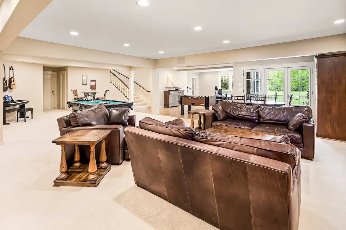 A wide view of a spacious downstairs family/recreation room showing two large brown leather sofas, wood and stone side tables, a carpeted floor, and a billiards pool table visible in the background on the left. Sliding glass doors lead outside. [Season: Indoor] [Lighting: Soft, warm recessed ceiling lights] [Content: Recreation room, leather sofas, pool table, downstairs living]