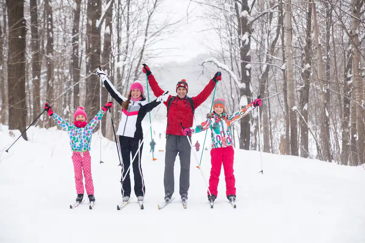 A joyful action photo of four guests (two adults, two kids) in colorful winter gear, raising their hands in excitement while cross-country skiing along a snow-covered forest path. [Season: Winter] [Lighting: Soft, diffused winter daylight] [Content: Cross-country skiing, winter forest, family activity, outdoor recreation]