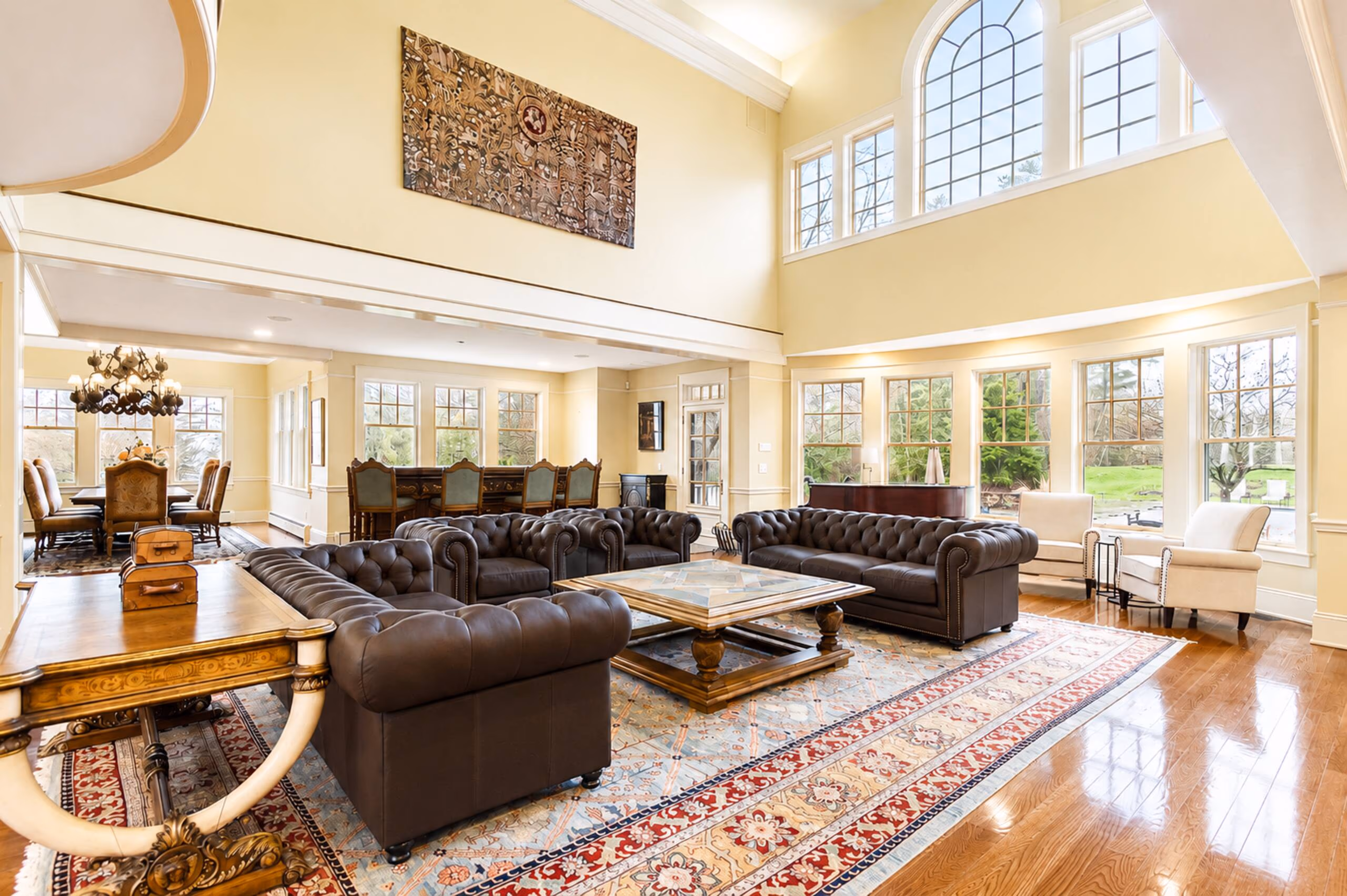 Panoramic, wide-angle interior view of the magnificent Great Room. Features soaring double-height ceilings, massive structural timber trusses, polished hardwood floors, two dark brown Chesterfield leather sofas, and a large red-patterned Persian rug. A mahogany grand piano sits in the corner, and a massive stone fireplace stands on the right. Large banks of windows look out on green lawns. [Season: Summer] [Lighting: Bright, natural daylight streaming in] [Content: Great room interior, timber trusses, Chesterfield sofas, grand piano, stone fireplace]