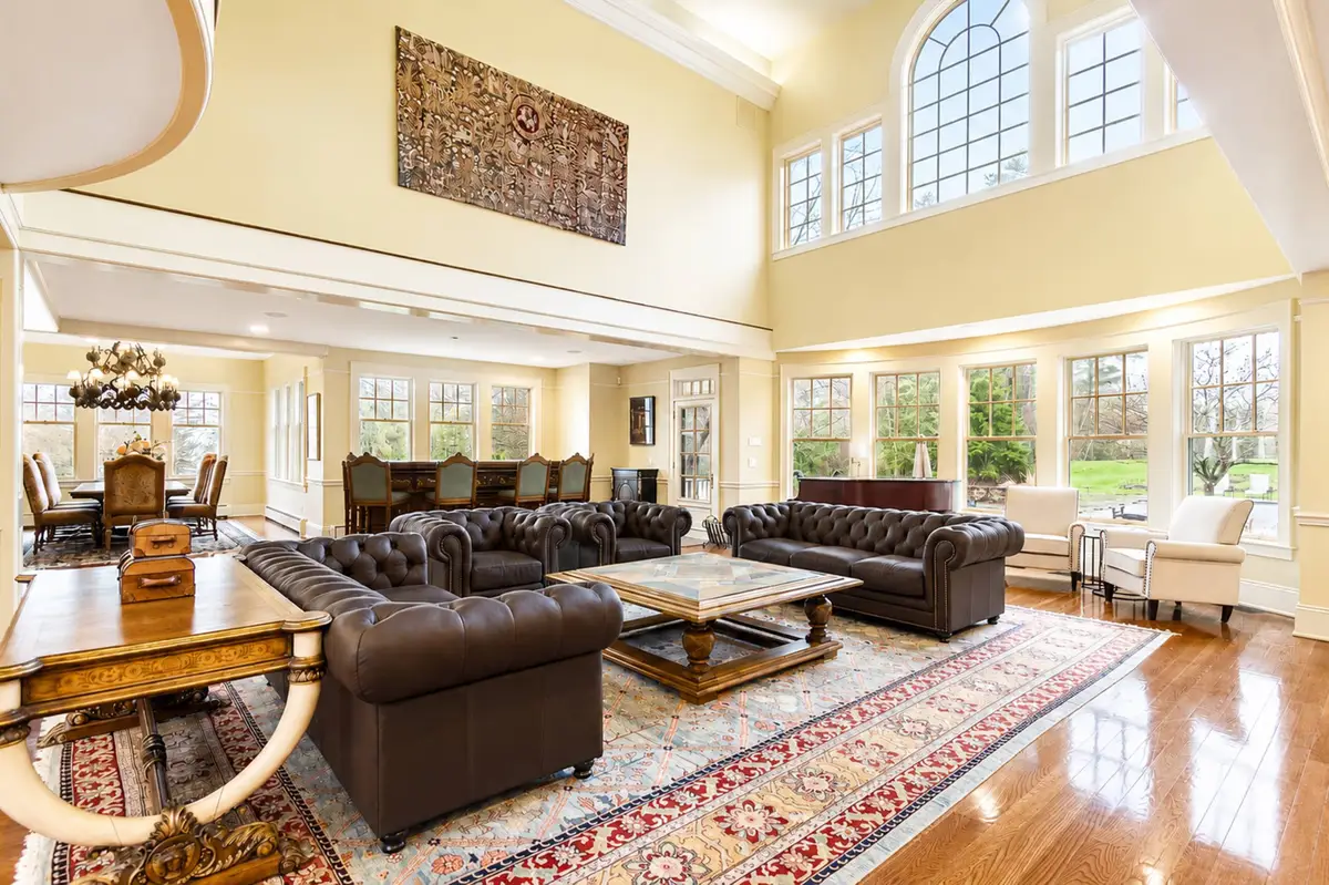 Panoramic, wide-angle interior view of the magnificent Great Room. Features soaring double-height ceilings, massive structural timber trusses, polished hardwood floors, two dark brown Chesterfield leather sofas, and a large red-patterned Persian rug. A mahogany grand piano sits in the corner, and a massive stone fireplace stands on the right. Large banks of windows look out on green lawns. [Season: Summer] [Lighting: Bright, natural daylight streaming in] [Content: Great room interior, timber trusses, Chesterfield sofas, grand piano, stone fireplace]