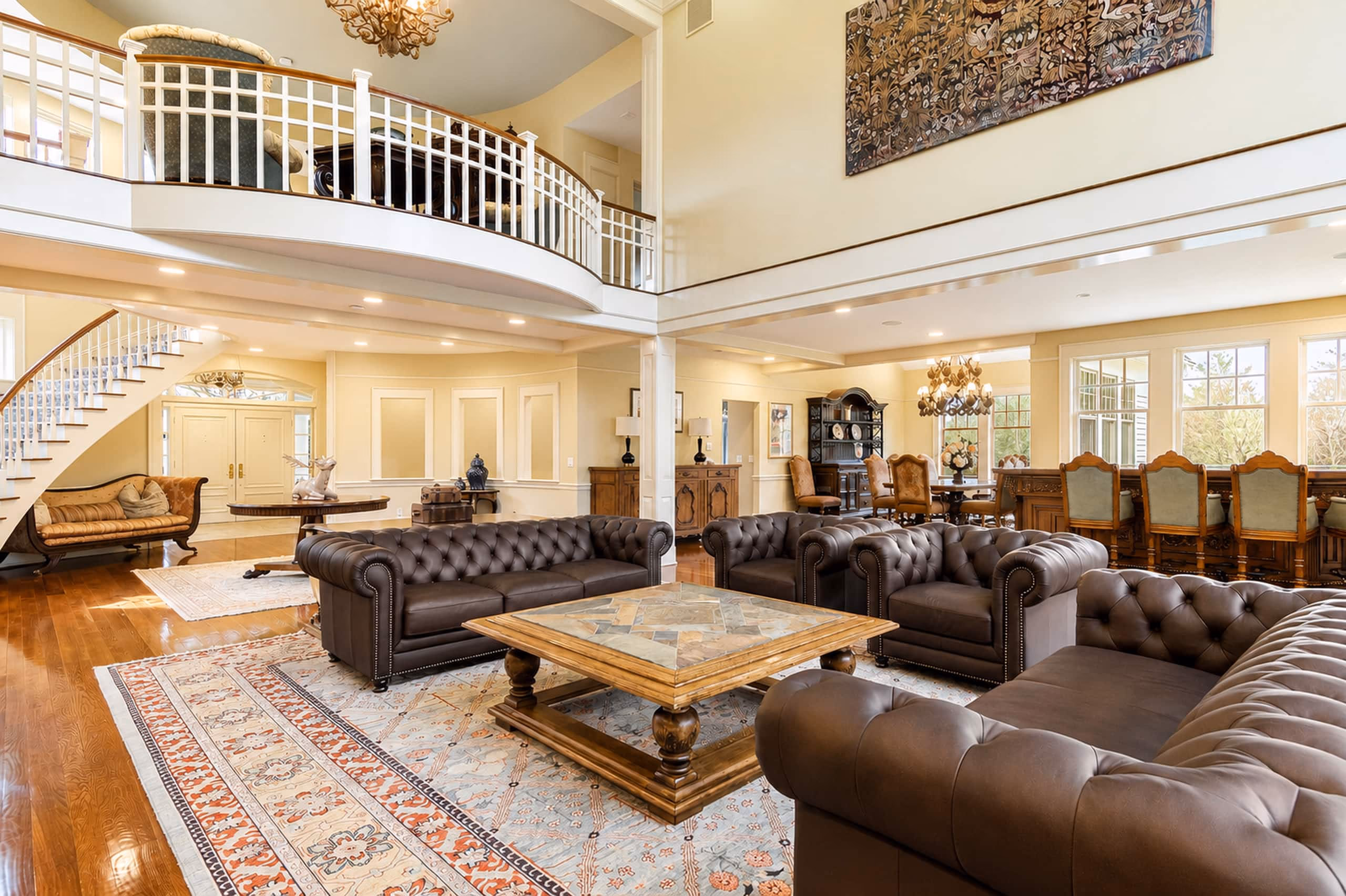 Another panoramic view of the Great Room from a slightly different angle, showing the two Chesterfield sofas, a heavy wooden coffee table with a stone inlay, a staircase and upper-level loft walkway in the background, and a formal dining table visible in the distance. [Season: Summer] [Lighting: Bright, natural daylight and warm ambient light] [Content: Great room interior, Chesterfield sofas, loft walkway, wooden stairs]