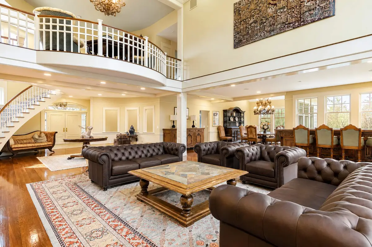 Another panoramic view of the Great Room from a slightly different angle, showing the two Chesterfield sofas, a heavy wooden coffee table with a stone inlay, a staircase and upper-level loft walkway in the background, and a formal dining table visible in the distance. [Season: Summer] [Lighting: Bright, natural daylight and warm ambient light] [Content: Great room interior, Chesterfield sofas, loft walkway, wooden stairs]