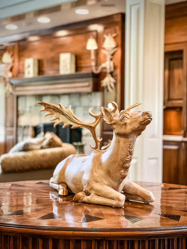 Close-up detail shot of an elegant wood-carved stag sculpture with gold-painted antlers, resting on a highly polished round wooden table featuring a geometric starburst inlay. The background shows a soft, warm out-of-focus view of the Great Room's stone fireplace, wood paneling, and glowing wall sconces. [Season: Autumn/Winter] [Lighting: Warm, focused indoor lighting] [Content: Sculpture close-up, wood table detail, fireplace background, decor]