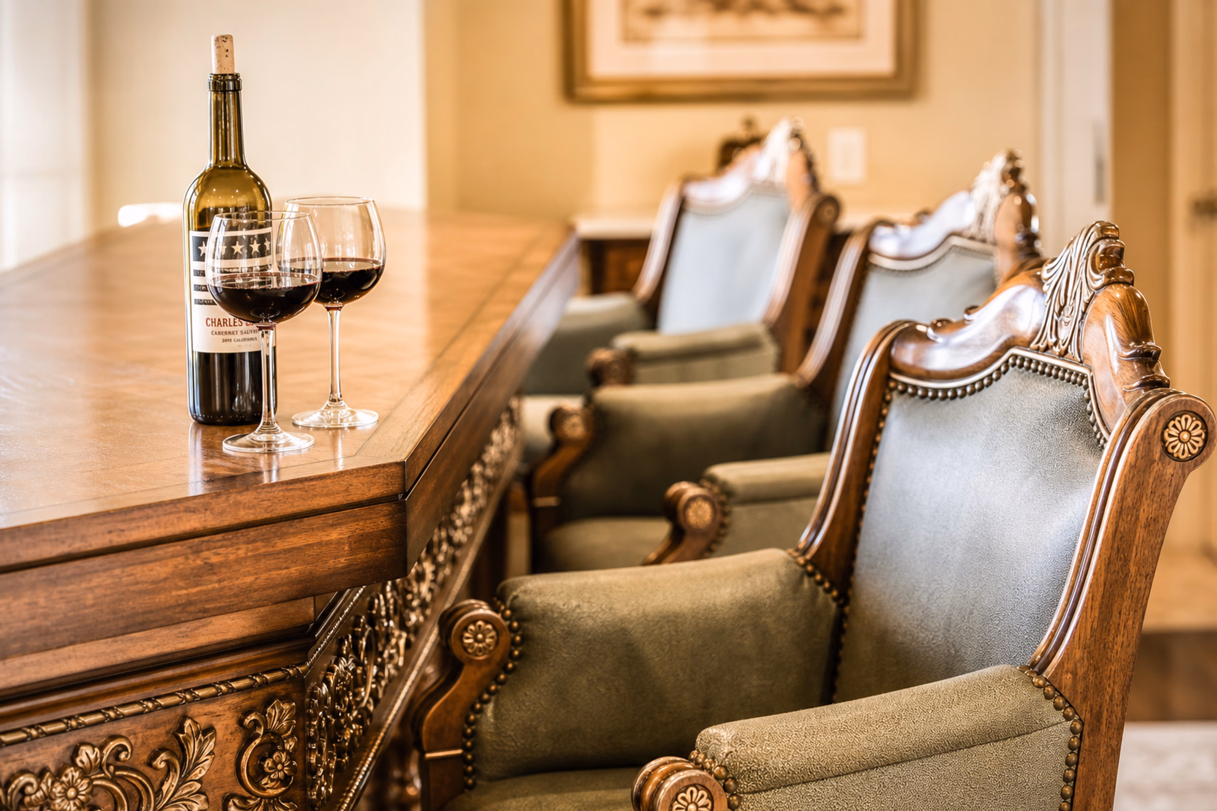 Close-up detail of a formal dining or conference table. Shows a long polished dark wood table set with a bottle of red wine and two crystal wine glasses, surrounded by high-backed classic chairs upholstered in premium antique sage green textured leather with brass nailhead trim. [Season: Autumn/Winter] [Lighting: Soft, warm ambient dining light] [Content: Dining table close-up, leather chairs, wine glasses, formal dining interior]