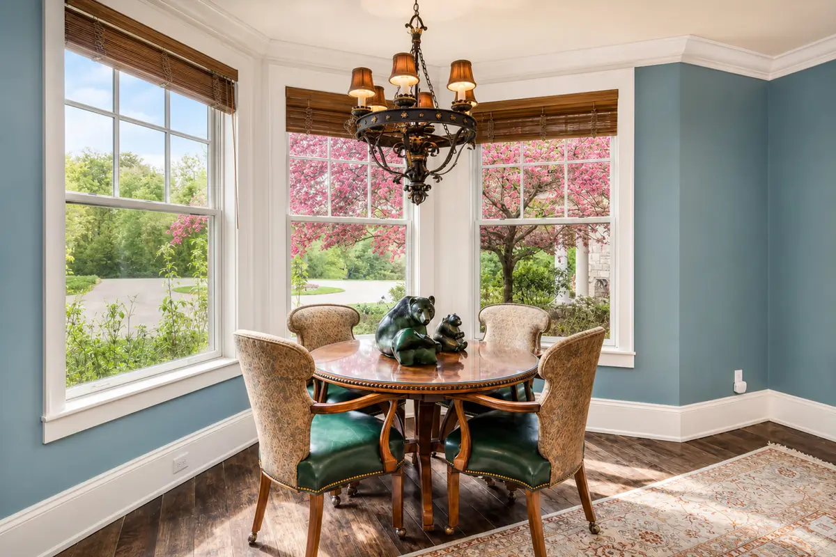 Brightly lit casual dining nook inside a multi-windowed bay alcove. Features a round dark wood table, four upholstered chairs with high backs, and an ornate dark metal chandelier. Large windows look out on a green lawn and pink blossoming trees. [Season: Spring] [Lighting: Bright, fresh morning daylight] [Content: Breakfast nook, dining table, bay window, spring garden exterior]
