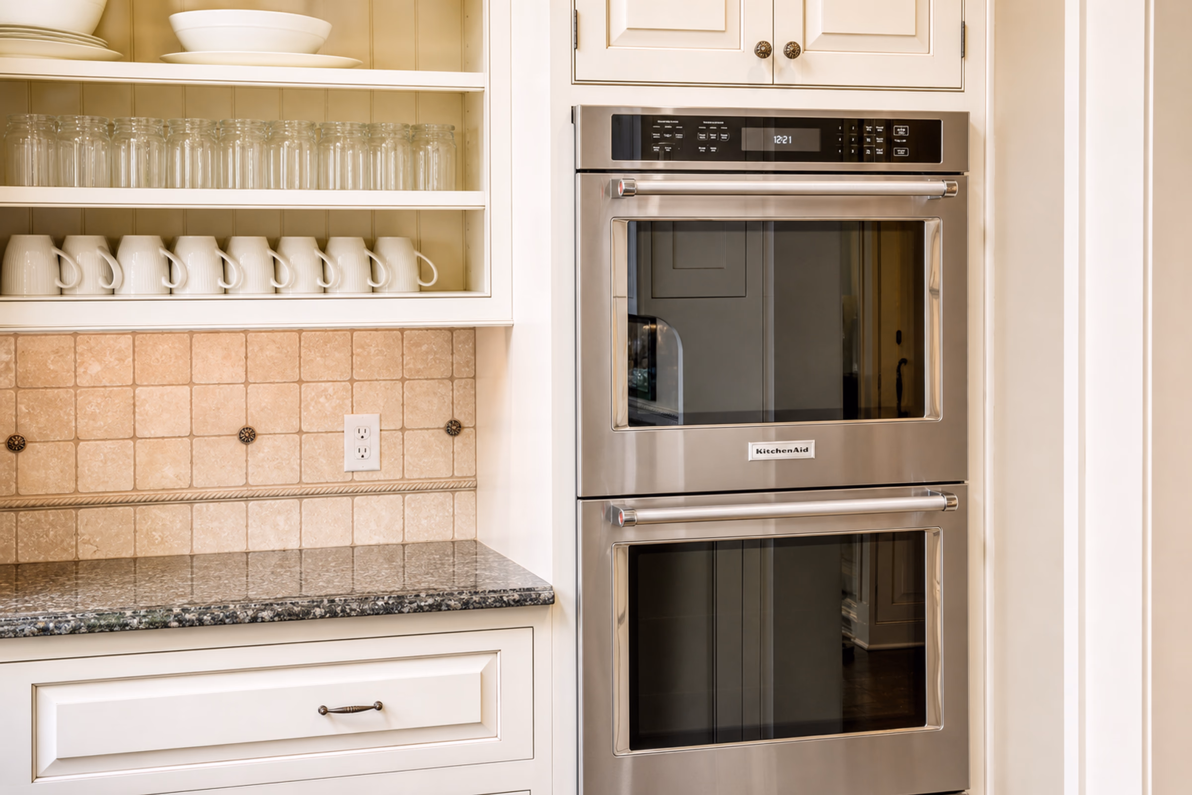Close-up of double built-in stainless steel professional wall ovens integrated into white cabinetry. To the left is a granite countertop, tile backsplash, and a shelf with white mugs. [Season: Indoor] [Lighting: Bright, clean kitchen lighting] [Content: Built-in ovens, stainless steel appliances, kitchen details]