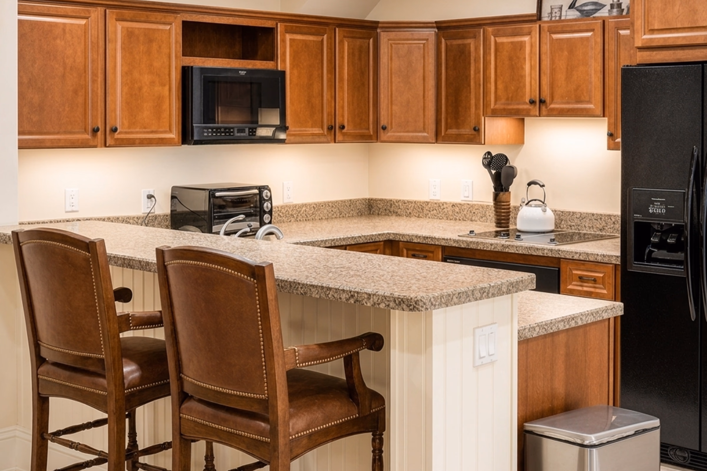 Secondary or prep kitchen showing warm wood cabinets, light-colored granite countertops, a peninsula bar with two matching leather barstool chairs, a stainless steel microwave and toaster oven, and a black refrigerator. [Season: Indoor] [Lighting: Bright, clear indoor light] [Content: Prep kitchen, wood cabinets, bar counter, appliances]