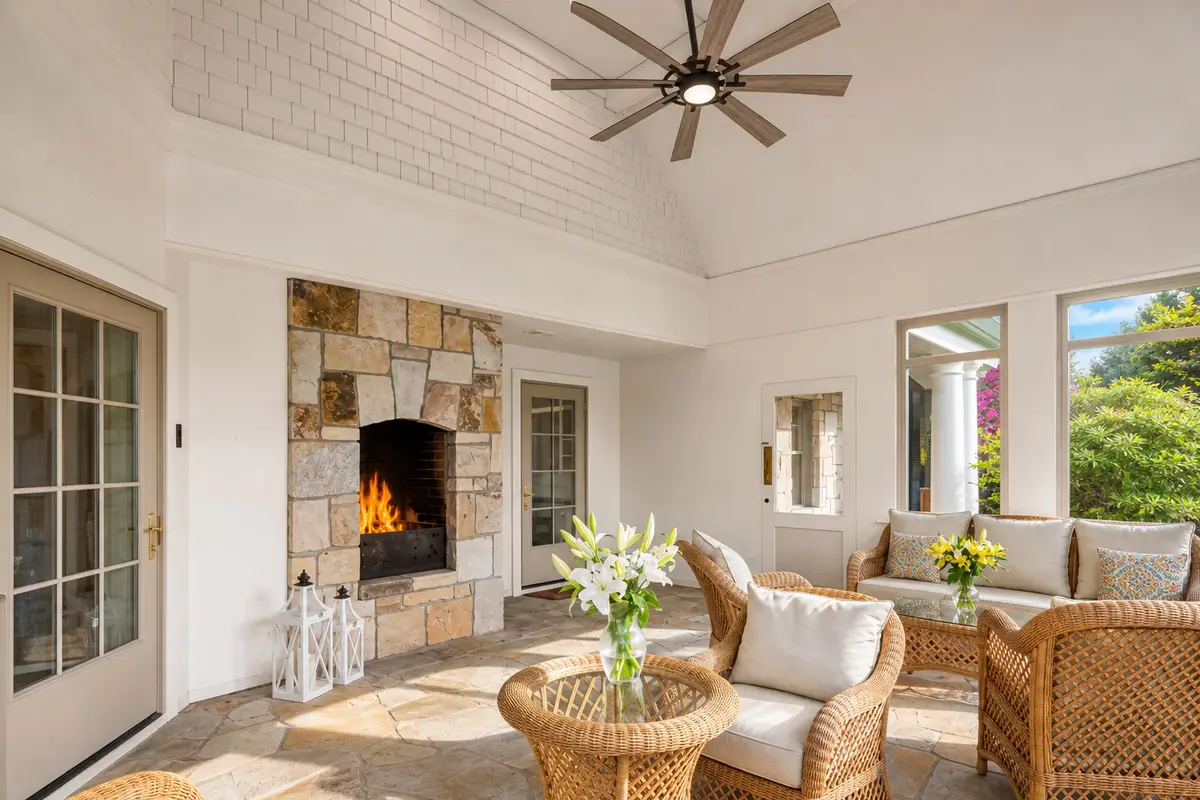 Another view of the bright sunroom showing the wicker seating, a large stone fireplace on the left with a lit fire, and white brick chimney walls. Large windows let in plenty of daylight. [Season: Summer/Spring] [Lighting: Bright daylight with warm fireplace glow] [Content: Sunroom, brick fireplace, wicker sofas, indoor-outdoor living]