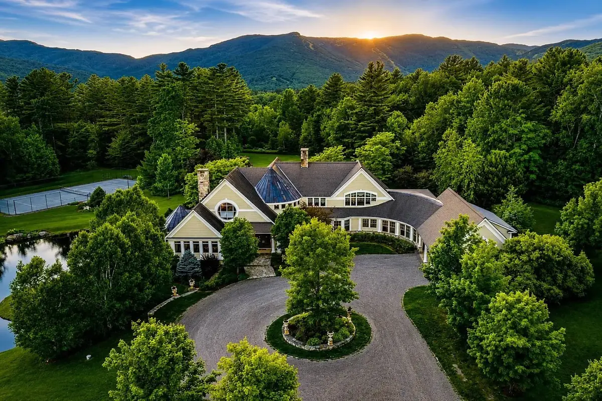 High-angle aerial view of the estate's front approach at dusk. Shows the circular gravel driveway with a central landscaped roundabout containing a mature tree, the front facade of the light-beige house with circular window accents, and a tennis court in the background, set against soft yellow-orange twilight mountains and green forests. [Season: Summer] [Lighting: Warm twilight, golden hour] [Content: Aerial view, front entrance, circular driveway, tennis court, landscape exterior]