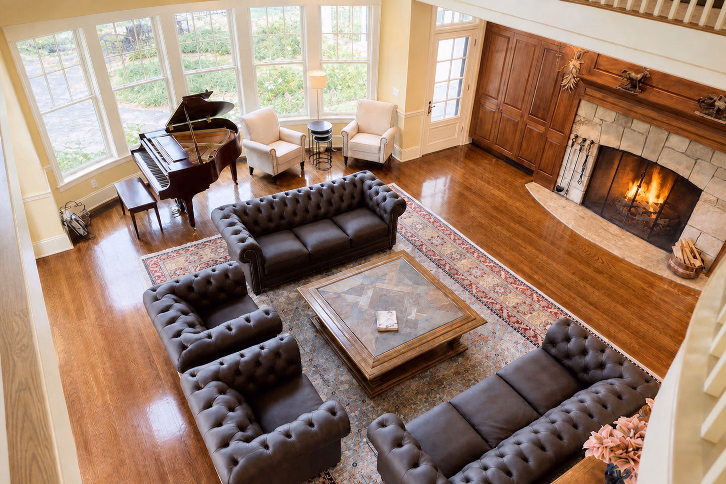 High-angle interior view looking down from the loft walkway onto the Great Room's spacious conversation seating area. Features two button-tufted grey Chesterfield sofas and two matching button-tufted armchairs arranged on a large red-patterned Persian rug around a square coffee table. A mahogany grand piano sits near large windows looking out at green gardens. Highlights polished hardwood floors and the edge of a massive stone fireplace. [Season: Summer] [Lighting: Bright, natural daytime light] [Content: Great room interior, furniture, piano, Persian rug, hardwood floors]