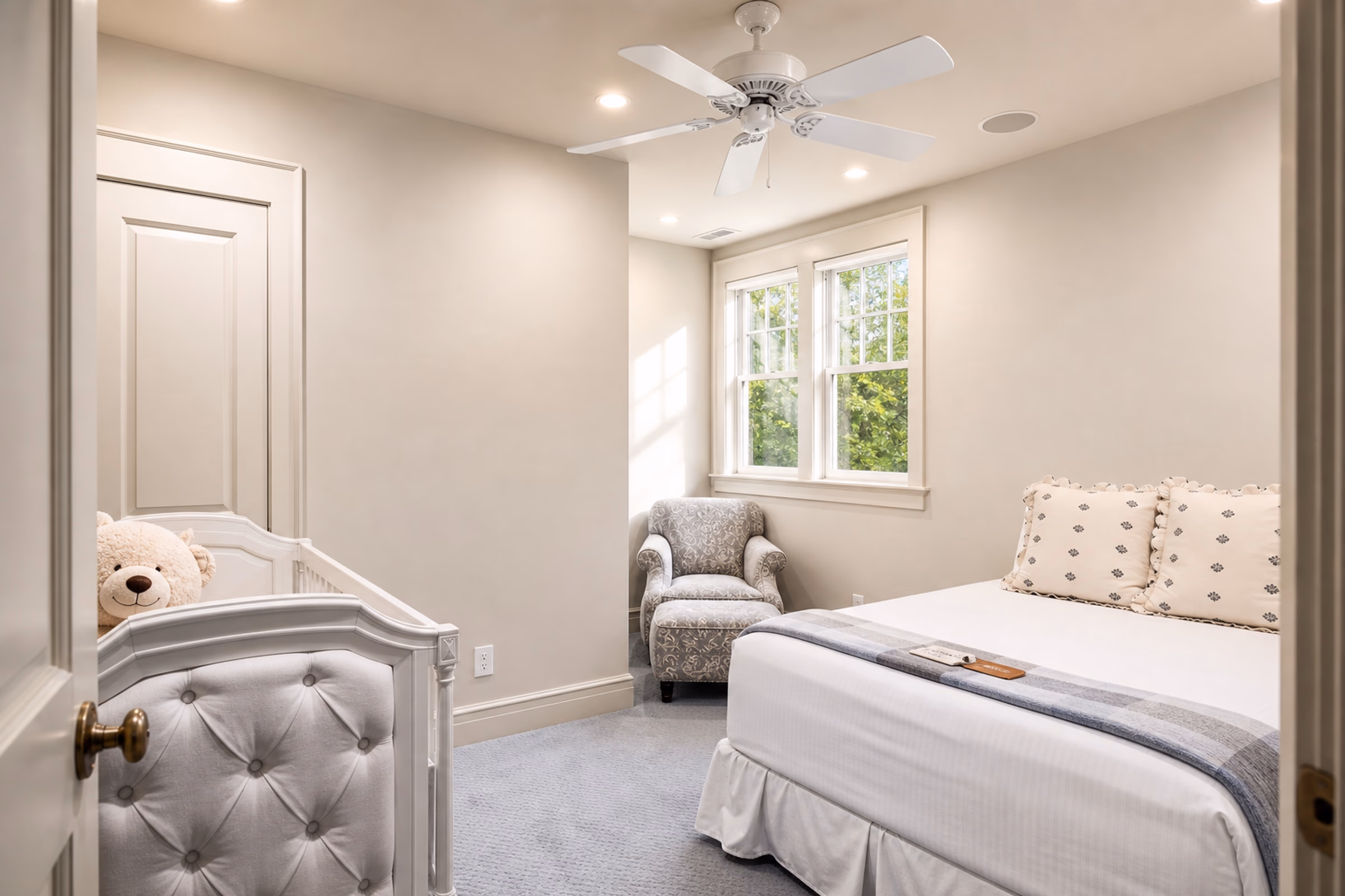Cozy guest bedroom featuring a queen bed with white linens, a ceiling fan, light beige walls, a grey upholstered armchair, and a window. [Season: Indoor] [Lighting: Soft, pleasant recessed ceiling lights] [Content: Cozy bedroom, queen bed, armchair, neutral tones]