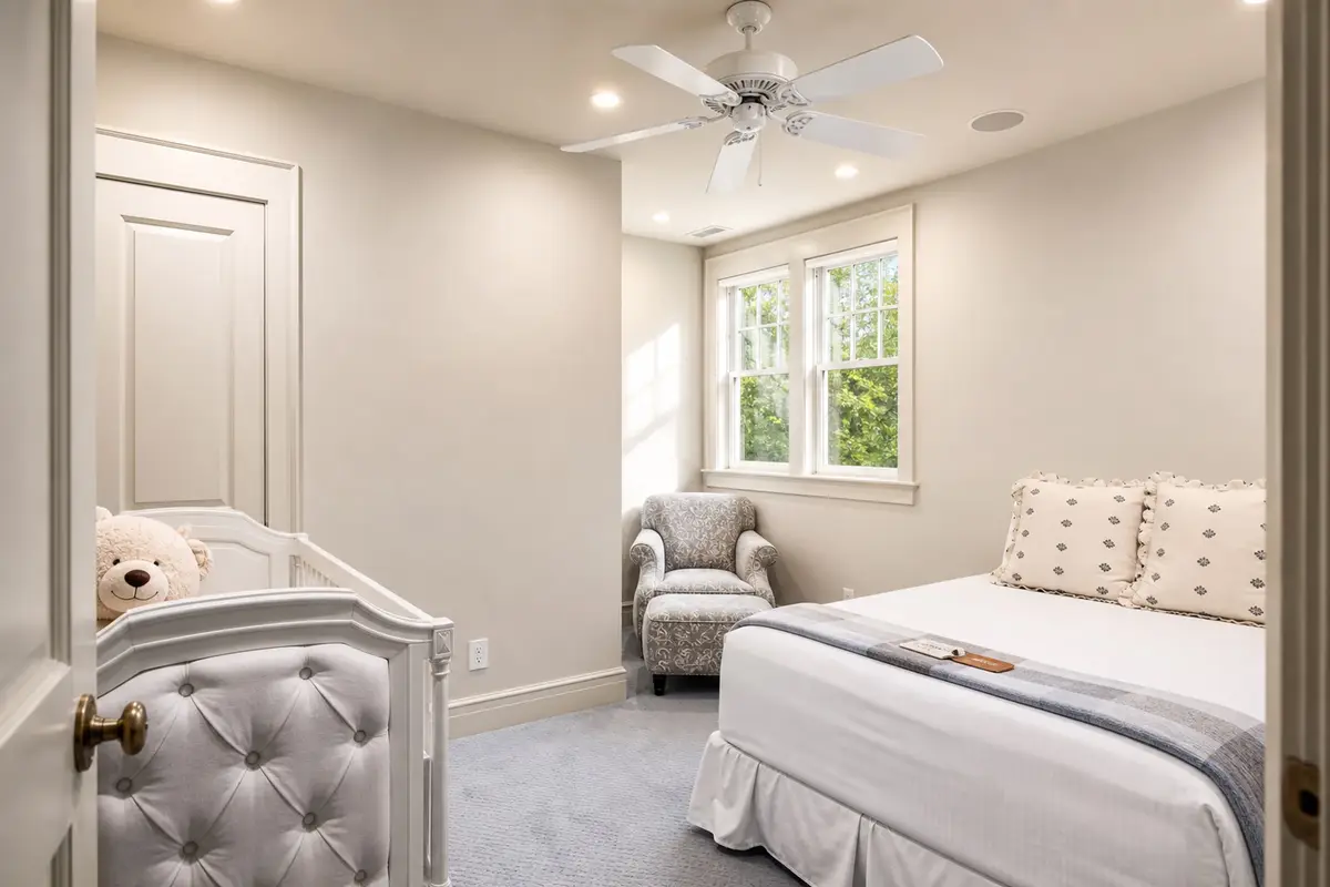 Cozy guest bedroom featuring a queen bed with white linens, a ceiling fan, light beige walls, a grey upholstered armchair, and a window. [Season: Indoor] [Lighting: Soft, pleasant recessed ceiling lights] [Content: Cozy bedroom, queen bed, armchair, neutral tones]